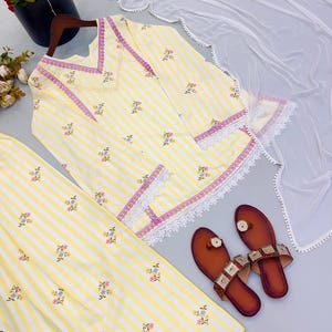 K&ouml;nnte beinhalten: Eine gelb-wei&szlig; gestreifte Tunika und eine passende Hose mit kleinen Blumenmustern, einem V-Ausschnitt mit violetter Borte, wei&szlig;er Spitzenverzierung an Saum und &Auml;rmeln, einem wei&szlig;en, transparenten Dupatta und einem Paar brauner flacher Sandalen.