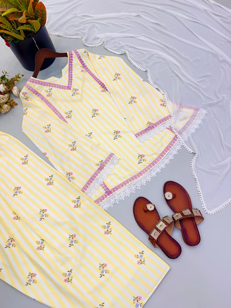 K&ouml;nnte beinhalten: Eine gelb-wei&szlig; gestreifte Tunika und eine passende Hose mit kleinen Blumenmustern, einem V-Ausschnitt mit violetter Borte, wei&szlig;er Spitzenverzierung an Saum und &Auml;rmeln, einem wei&szlig;en, transparenten Dupatta und einem Paar brauner flacher Sandalen.