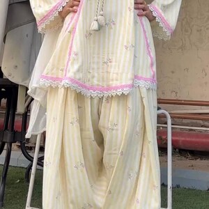K&ouml;nnte beinhalten: Ein hellgelb-wei&szlig; gestreiftes Tunika- und Dhoti-Hosen-Set mit kleinen Blumenmustern, rosa Spitzenbesatz an Ausschnitt, &Auml;rmeln und Saum sowie einer dekorativen Kordel mit Quasten, die mittig vom Ausschnitt herabh&auml;ngt.