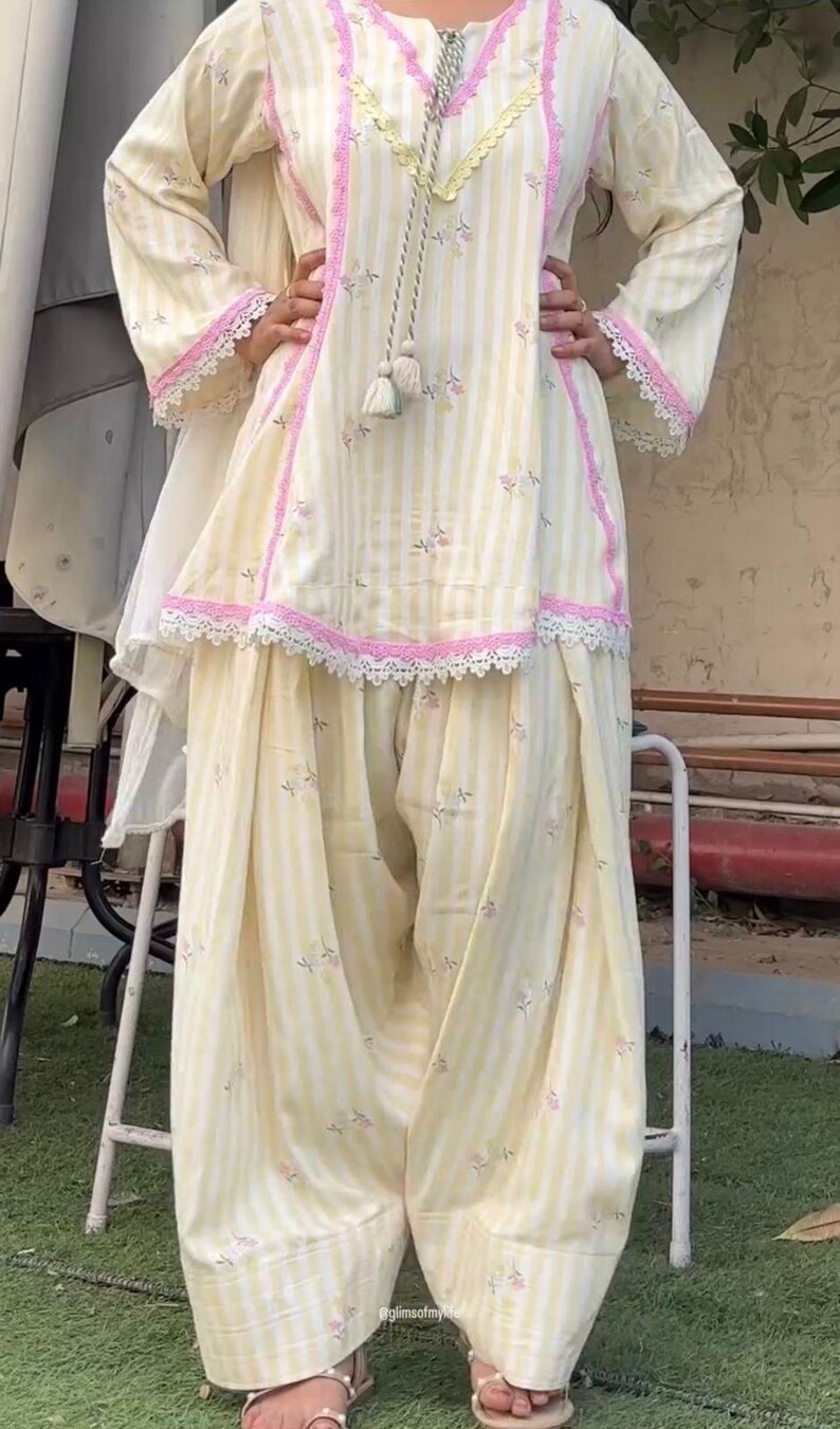 K&ouml;nnte beinhalten: Ein hellgelb-wei&szlig; gestreiftes Tunika- und Dhoti-Hosen-Set mit kleinen Blumenmustern, rosa Spitzenbesatz an Ausschnitt, &Auml;rmeln und Saum sowie einer dekorativen Kordel mit Quasten, die mittig vom Ausschnitt herabh&auml;ngt.