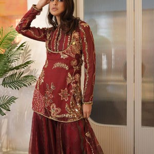 May include: Burgundy-colored formal wear with intricate gold embroidery. The long-sleeved top features floral and geometric patterns, embellished with sequins. The matching skirt has a flowing, layered design, creating an elegant silhouette. This ensemble is suitable for special occasions.
