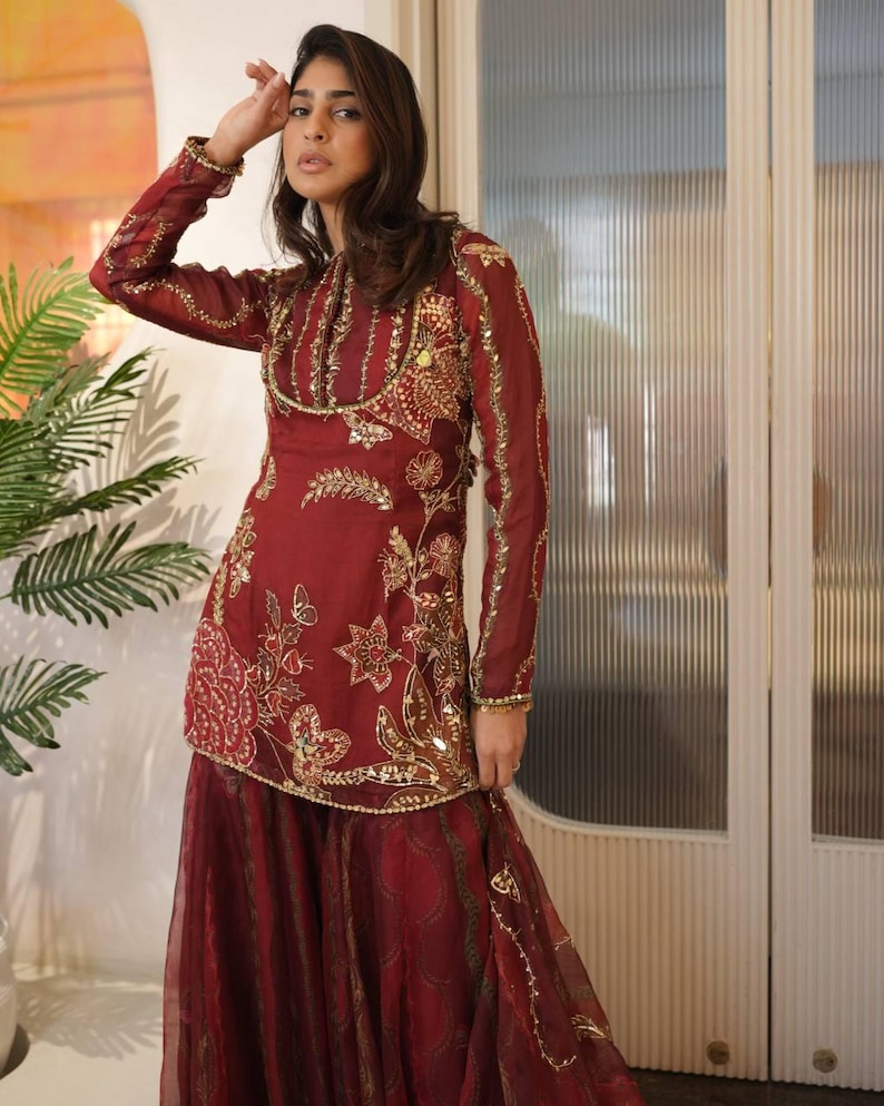 May include: Burgundy-colored formal wear with intricate gold embroidery. The long-sleeved top features floral and geometric patterns, embellished with sequins. The matching skirt has a flowing, layered design, creating an elegant silhouette. This ensemble is suitable for special occasions.