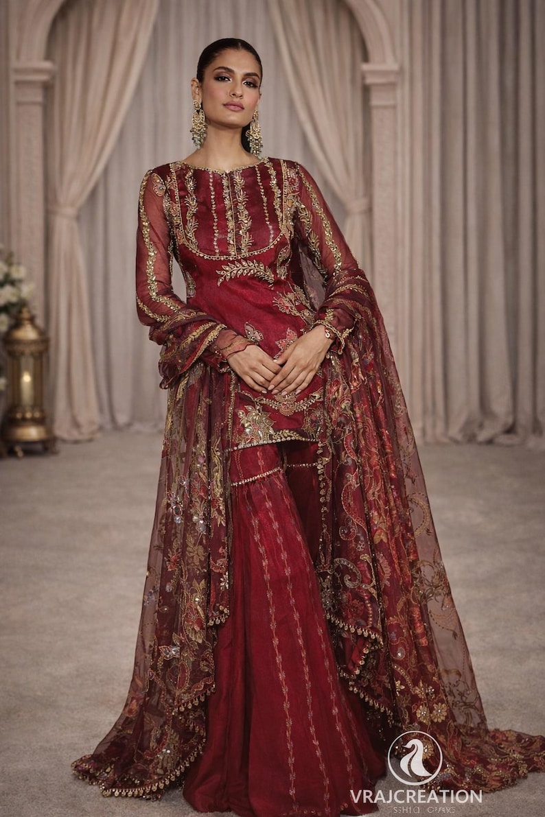 May include: A burgundy-colored, embroidered formal outfit. The ensemble includes a long-sleeved top, wide-leg pants, and a sheer shawl. Gold embroidery and embellishments adorn the outfit. The outfit is from Vrajcreation.