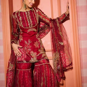 May include: A red embroidered ensemble featuring a long-sleeved tunic and wide-legged pants. The outfit is adorned with gold embellishments and floral patterns. The sheer dupatta adds a layer of elegance. The ensemble is a traditional garment.