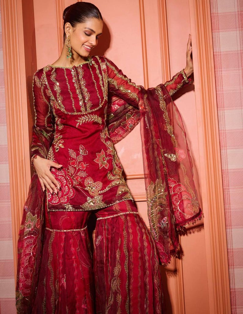 May include: A red embroidered ensemble featuring a long-sleeved tunic and wide-legged pants. The outfit is adorned with gold embellishments and floral patterns. The sheer dupatta adds a layer of elegance. The ensemble is a traditional garment.