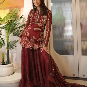 May include: Burgundy-colored formal outfit with gold embroidery. The long-sleeved top features intricate gold detailing around the neckline, sleeves, and bodice. The outfit includes a matching skirt with a flowing design. The ensemble is suitable for special occasions.