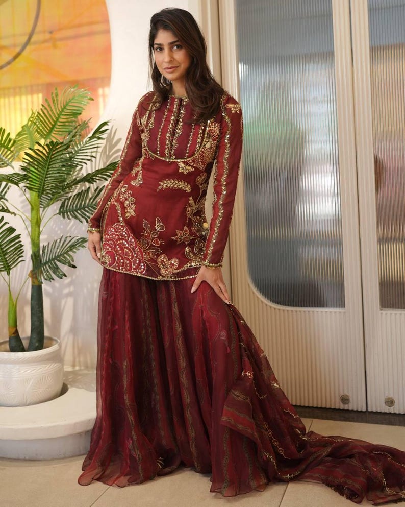 May include: Burgundy-colored formal outfit with gold embroidery. The long-sleeved top features intricate gold detailing around the neckline, sleeves, and bodice. The outfit includes a matching skirt with a flowing design. The ensemble is suitable for special occasions.