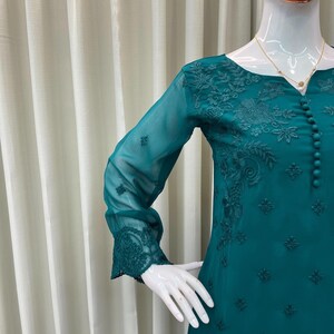 May include: A teal long-sleeved tunic featuring intricate tonal floral embroidery, a round neckline, and a vertical row of small matching fabric buttons down the center front. The sheer sleeves are finished with detailed embroidered cuffs.
