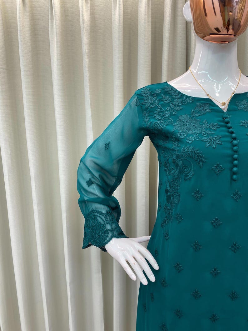 May include: A teal long-sleeved tunic featuring intricate tonal floral embroidery, a round neckline, and a vertical row of small matching fabric buttons down the center front. The sheer sleeves are finished with detailed embroidered cuffs.