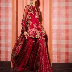May include: A vibrant red ensemble featuring a tunic and wide-leg pants, adorned with intricate gold embroidery. The outfit includes a matching sheer scarf. The tunic has long sleeves and a round neckline. The pants have a flowing, layered design.