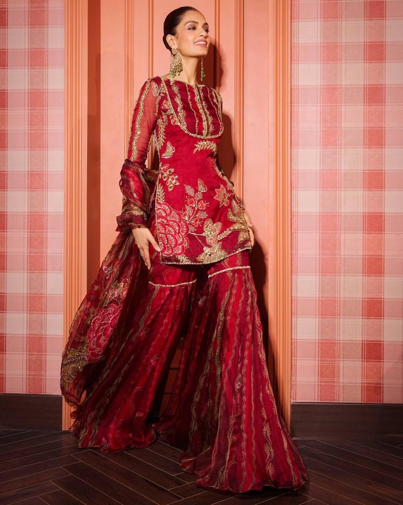 May include: A vibrant red ensemble featuring a tunic and wide-leg pants, adorned with intricate gold embroidery. The outfit includes a matching sheer scarf. The tunic has long sleeves and a round neckline. The pants have a flowing, layered design.