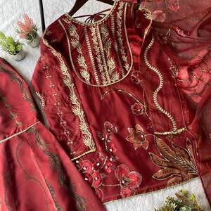 May include: A maroon embroidered outfit, including a top and skirt, with gold accents. The top features intricate floral embroidery and a detailed neckline. The skirt has a matching pattern. A sheer maroon scarf is also included.
