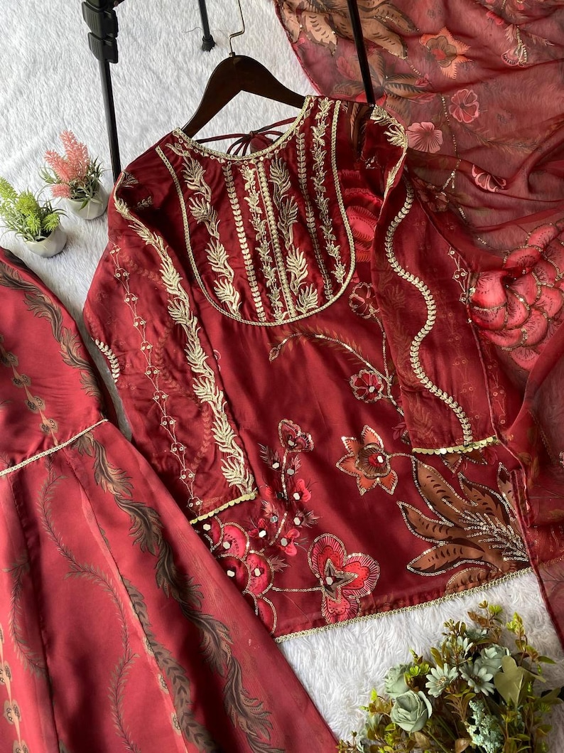 May include: A maroon embroidered outfit, including a top and skirt, with gold accents. The top features intricate floral embroidery and a detailed neckline. The skirt has a matching pattern. A sheer maroon scarf is also included.