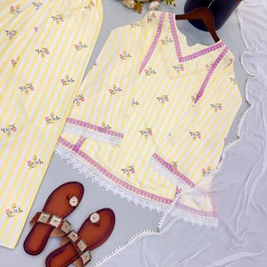 K&ouml;nnte beinhalten: Ein gelb-wei&szlig; gestreiftes Tunika- und Hosenset mit kleiner Blumenstickerei, lila Besatz und wei&szlig;er Spitzenverzierung an Saum und &Auml;rmeln, dazu ein Paar braune Sandalen und ein wei&szlig;er, transparenter Dupatta.