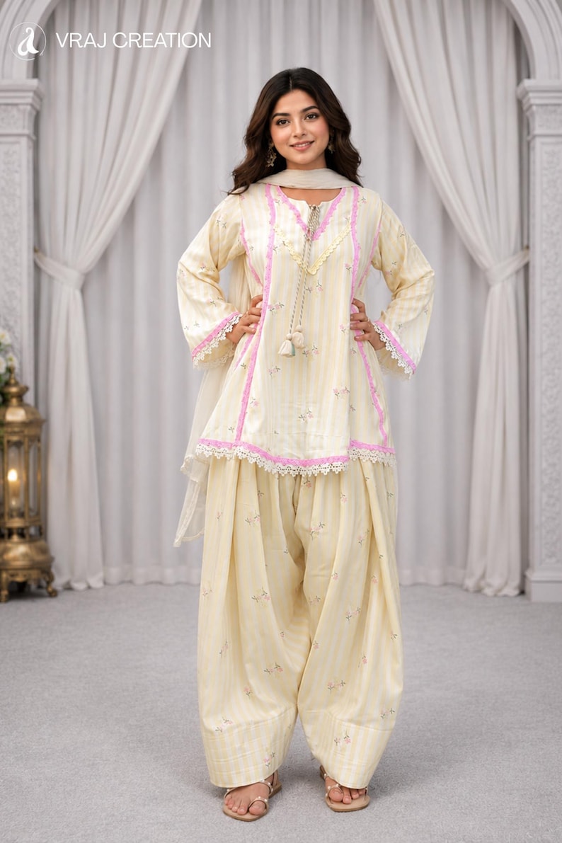 K&ouml;nnte beinhalten: Ein cremefarbenes Salwar-Kameez-Set bestehend aus einer Tunika mit vertikalen Streifen, rosa Spitzenbesatz und einem Schn&uuml;rhalsausschnitt mit Quasten. Die passende weite Hose und der transparente Dupatta sind mit einem zarten Blumenmuster verziert.