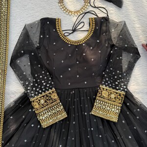 Peut inclure: Robe de soir&eacute;e noire avec de longues manches transparentes et des broderies dor&eacute;es aux poignets. La robe pr&eacute;sente une encolure ronde avec une bordure dor&eacute;e et est orn&eacute;e de paillettes argent&eacute;es. Un collier dor&eacute; et des accents de tulle noir sont &eacute;galement visibles.