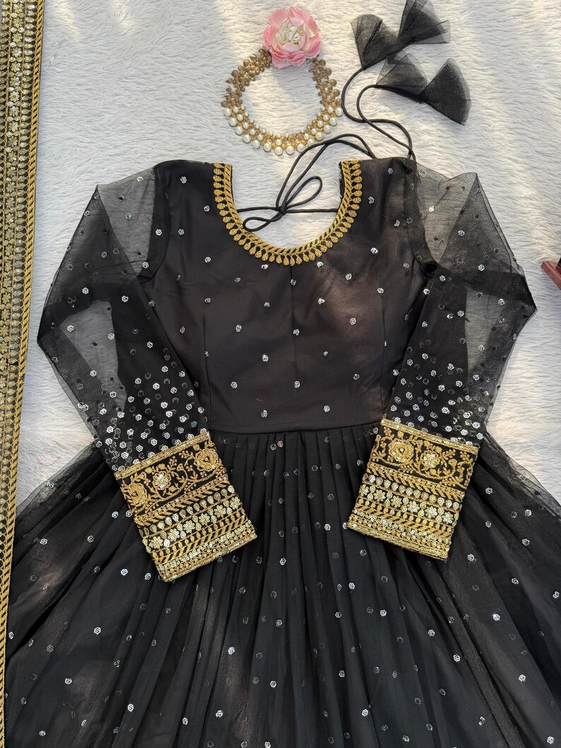 Peut inclure: Robe de soir&eacute;e noire avec de longues manches transparentes et des broderies dor&eacute;es aux poignets. La robe pr&eacute;sente une encolure ronde avec une bordure dor&eacute;e et est orn&eacute;e de paillettes argent&eacute;es. Un collier dor&eacute; et des accents de tulle noir sont &eacute;galement visibles.
