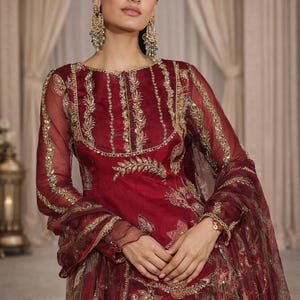 May include: A burgundy-colored formal dress with gold embroidery. The dress features long sleeves and intricate floral patterns. The image also shows matching earrings and a bracelet. The text "VRAJCREATION" is visible at the bottom.