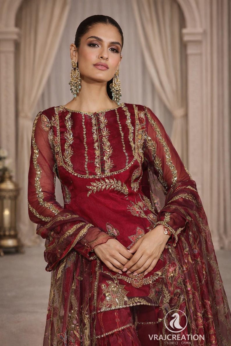 May include: A burgundy-colored formal dress with gold embroidery. The dress features long sleeves and intricate floral patterns. The image also shows matching earrings and a bracelet. The text "VRAJCREATION" is visible at the bottom.