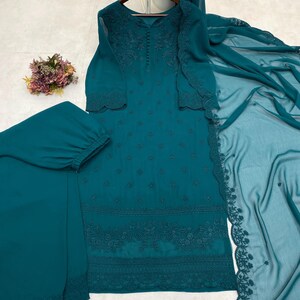 May include: A teal long-sleeved tunic with intricate tonal embroidery and a row of small buttons at the neckline. The set includes matching teal trousers with a cuffed hem and a sheer, embroidered teal dupatta draped to the side.