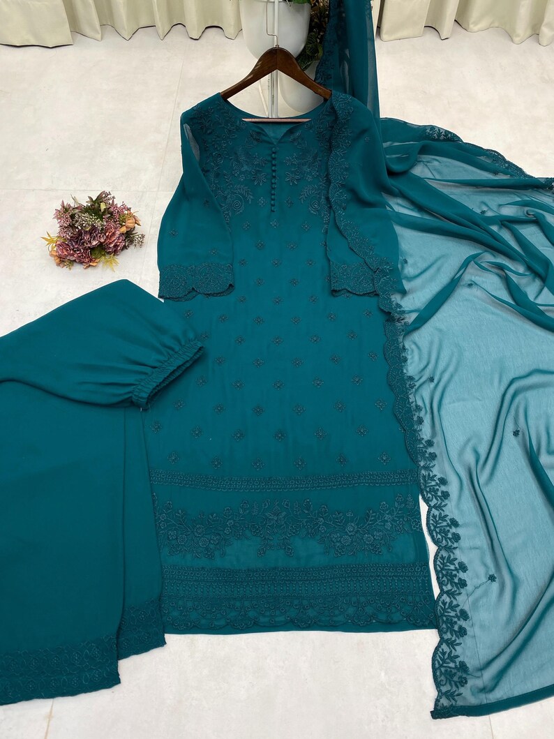 May include: A teal long-sleeved tunic with intricate tonal embroidery and a row of small buttons at the neckline. The set includes matching teal trousers with a cuffed hem and a sheer, embroidered teal dupatta draped to the side.