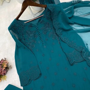 May include: A teal long-sleeved tunic featuring intricate tonal embroidery, a row of small fabric-covered buttons down the center chest, and scalloped cuffs. The garment is made of a sheer, lightweight fabric and includes a matching sheer scarf.