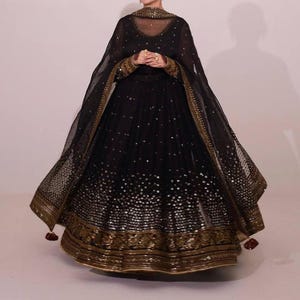 Peut inclure: Une robe de soir&eacute;e noire orn&eacute;e de d&eacute;tails dor&eacute;s. La robe pr&eacute;sente un d&eacute;collet&eacute; et des manches transparents, avec des broderies dor&eacute;es complexes sur les poignets et l'ourlet. La jupe ample est orn&eacute;e de paillettes &eacute;parses. Un ch&acirc;le transparent assorti compl&egrave;te l'ensemble.