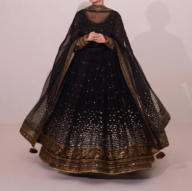 Peut inclure: Une robe de soir&eacute;e noire orn&eacute;e de d&eacute;tails dor&eacute;s. La robe pr&eacute;sente un d&eacute;collet&eacute; et des manches transparents, avec des broderies dor&eacute;es complexes sur les poignets et l'ourlet. La jupe ample est orn&eacute;e de paillettes &eacute;parses. Un ch&acirc;le transparent assorti compl&egrave;te l'ensemble.