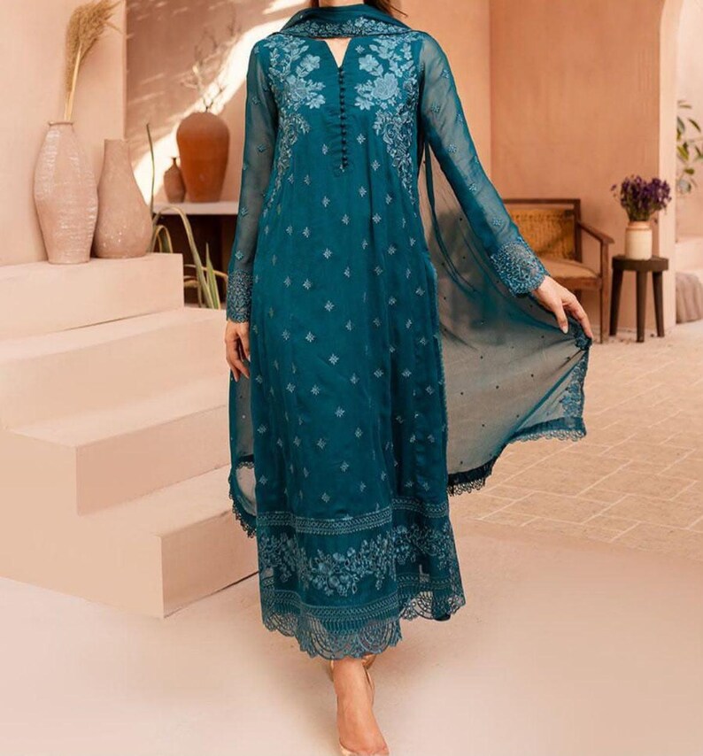 May include: A long-sleeved teal dress featuring intricate tonal floral embroidery on the bodice and hem, a notched neckline with a vertical row of small buttons, and a sheer, flowing overlay that extends into a draped panel on the side.