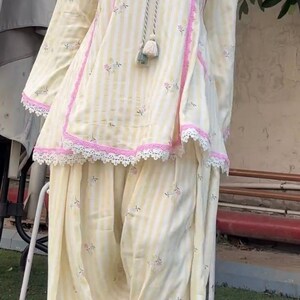 K&ouml;nnte beinhalten: Ein hellgelb-wei&szlig; gestreiftes Tunika- und Hosen-Set mit kleinen Blumenmustern. Die Tunika hat eine rosa Verzierung, wei&szlig;e Spitzenkanten am Saum und eine dekorative Kordel mit Quasten am Ausschnitt. Ein wei&szlig;er Schal ist ebenfalls Teil des Outfits.