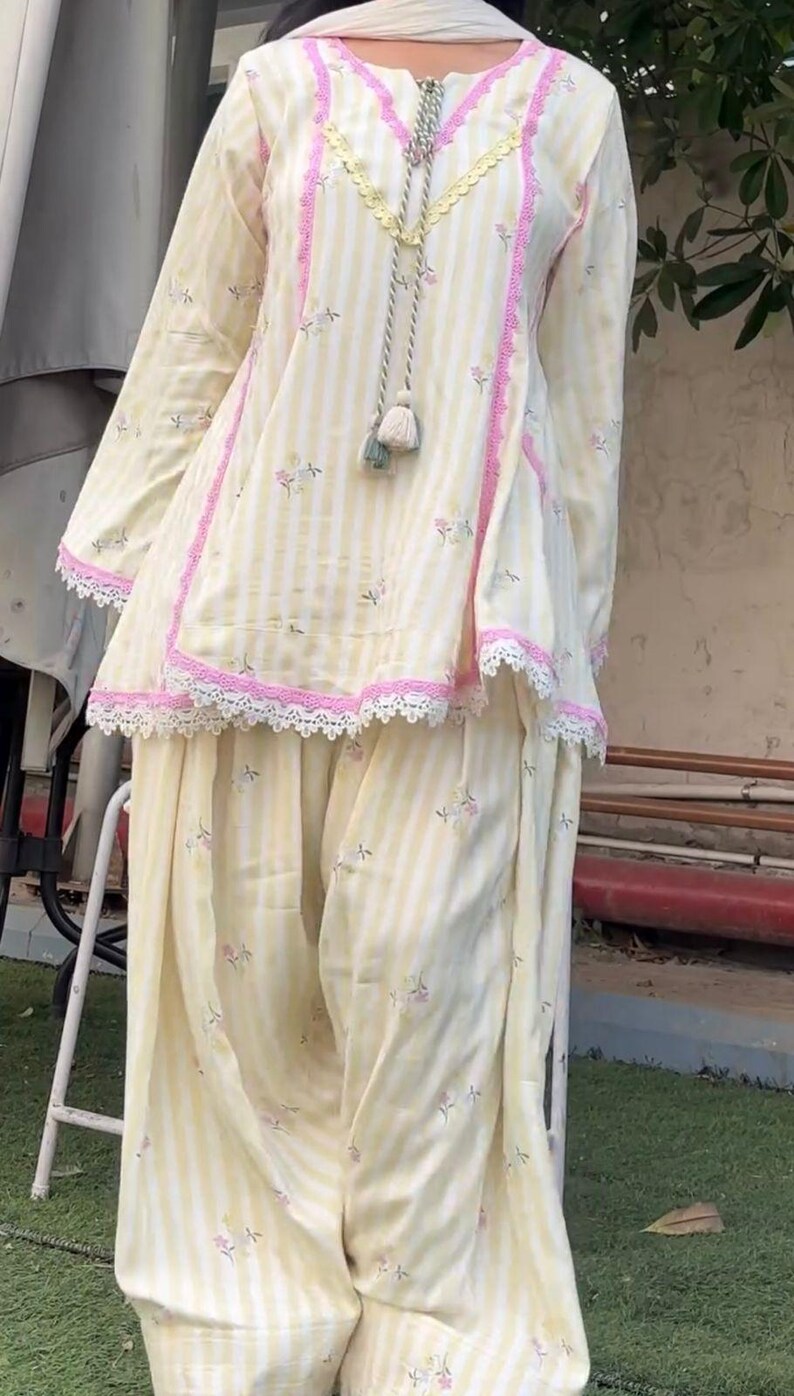 K&ouml;nnte beinhalten: Ein hellgelb-wei&szlig; gestreiftes Tunika- und Hosen-Set mit kleinen Blumenmustern. Die Tunika hat eine rosa Verzierung, wei&szlig;e Spitzenkanten am Saum und eine dekorative Kordel mit Quasten am Ausschnitt. Ein wei&szlig;er Schal ist ebenfalls Teil des Outfits.