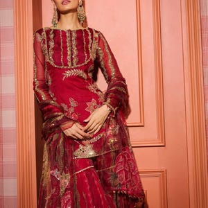 May include: A red embroidered outfit with gold accents. The ensemble includes a long-sleeved top, matching pants, and a scarf. The outfit features intricate floral and geometric patterns, with embellishments around the neckline and sleeves. The fabric appears sheer and flowing.