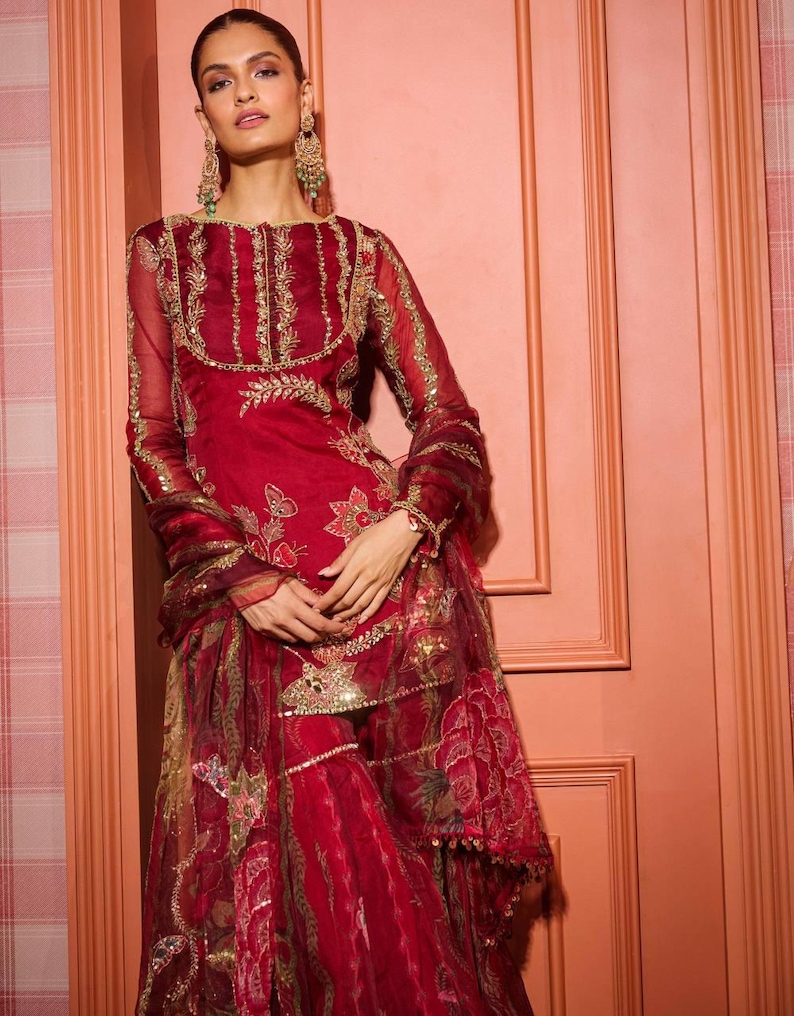 May include: A red embroidered outfit with gold accents. The ensemble includes a long-sleeved top, matching pants, and a scarf. The outfit features intricate floral and geometric patterns, with embellishments around the neckline and sleeves. The fabric appears sheer and flowing.
