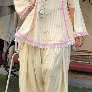 K&ouml;nnte beinhalten: Ein zweiteiliges Outfit bestehend aus einer hellgelb-wei&szlig; gestreiften Tunika mit kleinen Blumenakzenten, rosa Spitzenbesatz und einem dekorativen Quasten-Kordelzug am Ausschnitt, kombiniert mit einer passenden, locker sitzenden Plisseehose.