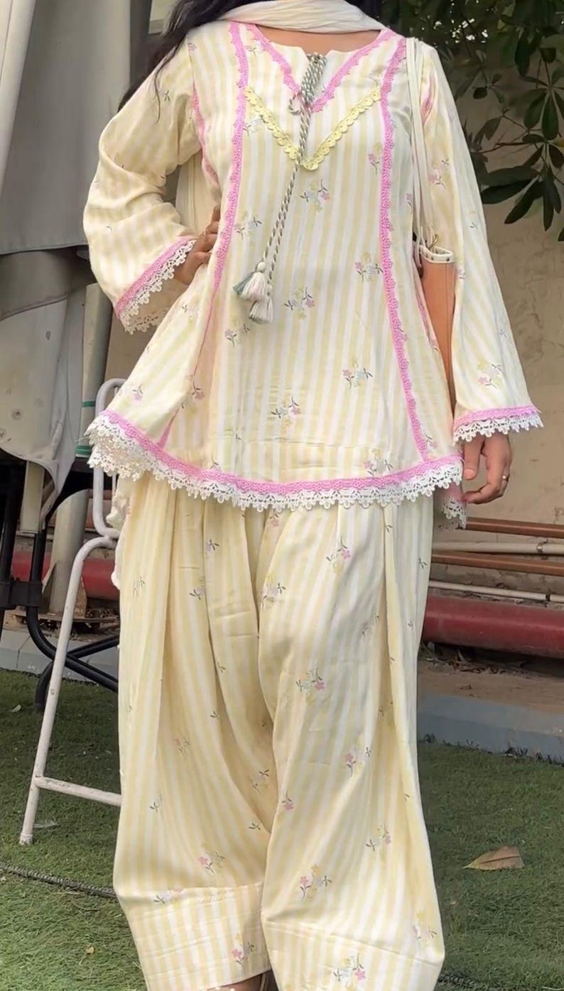 K&ouml;nnte beinhalten: Ein zweiteiliges Outfit bestehend aus einer hellgelb-wei&szlig; gestreiften Tunika mit kleinen Blumenakzenten, rosa Spitzenbesatz und einem dekorativen Quasten-Kordelzug am Ausschnitt, kombiniert mit einer passenden, locker sitzenden Plisseehose.