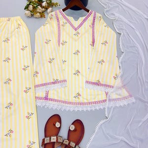 K&ouml;nnte beinhalten: Eine gelb-wei&szlig; gestreifte Tunika mit Blumenstickerei, passender Hose und einem wei&szlig;en, transparenten Dupatta mit Bommelborte. Die Tunika hat einen V-Ausschnitt mit lila Spitzenbesatz sowie wei&szlig;e Spitzenborten am Saum und an den &Auml;rmeln.