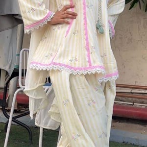 K&ouml;nnte beinhalten: Ein Set aus einer Tunika und einer Hose mit hellgelben und wei&szlig;en Streifen sowie kleinen Blumenmustern. Die Tunika hat lange &Auml;rmel, einen V-Ausschnitt mit dekorativen Quasten und eine rosa Spitzenborte entlang der N&auml;hte und am Saum.