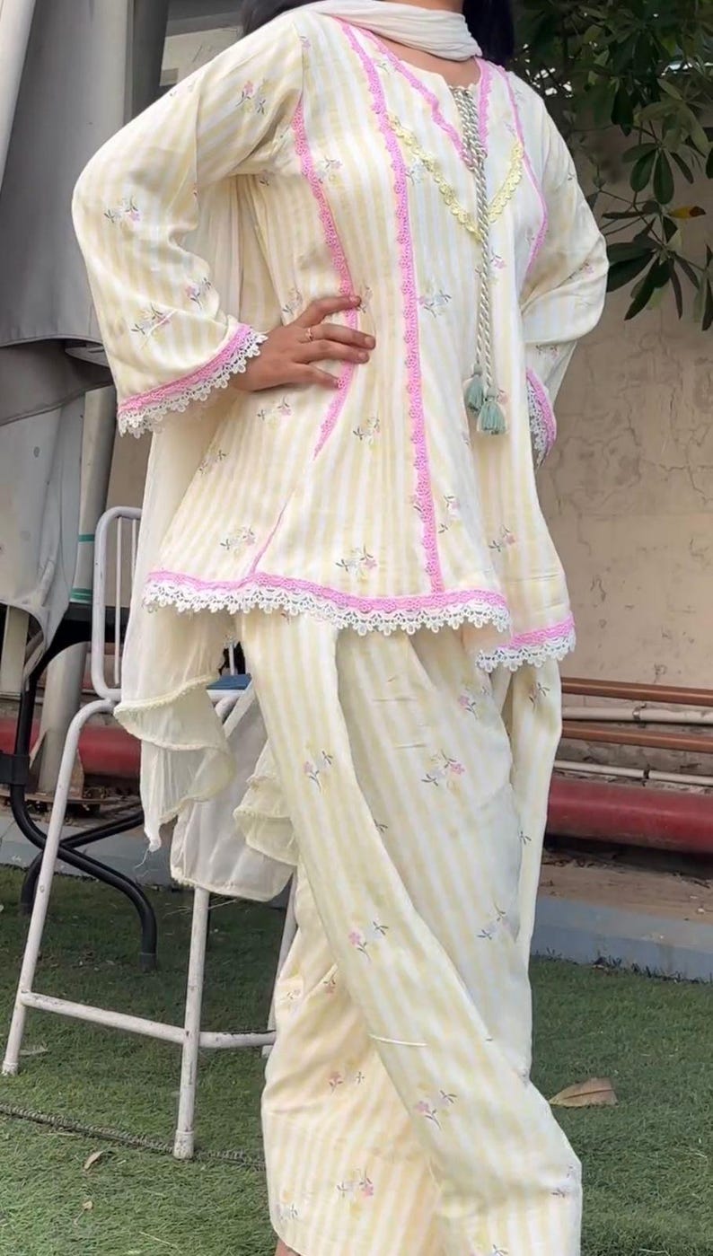K&ouml;nnte beinhalten: Ein Set aus einer Tunika und einer Hose mit hellgelben und wei&szlig;en Streifen sowie kleinen Blumenmustern. Die Tunika hat lange &Auml;rmel, einen V-Ausschnitt mit dekorativen Quasten und eine rosa Spitzenborte entlang der N&auml;hte und am Saum.