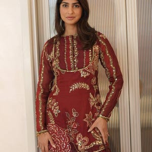 May include: A burgundy-colored, long-sleeved tunic and skirt set. The tunic features intricate gold embroidery along the neckline, sleeves, and bodice, with floral and geometric patterns. The skirt complements the tunic with a matching color and design.