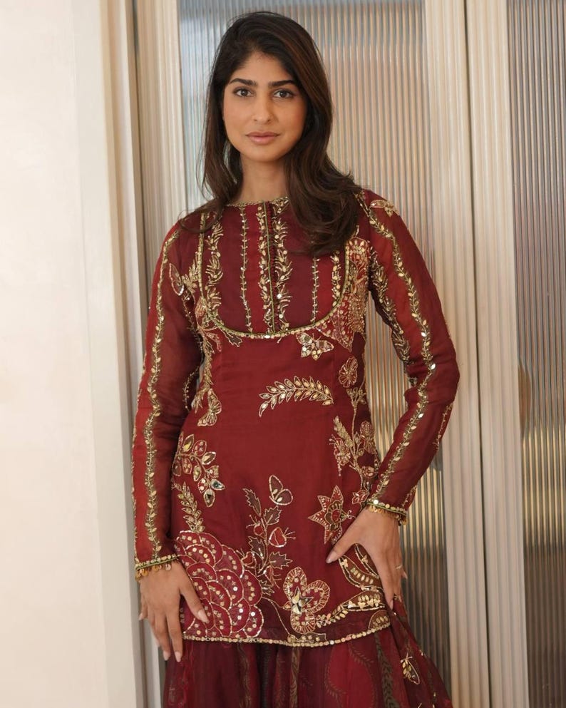 May include: A burgundy-colored, long-sleeved tunic and skirt set. The tunic features intricate gold embroidery along the neckline, sleeves, and bodice, with floral and geometric patterns. The skirt complements the tunic with a matching color and design.