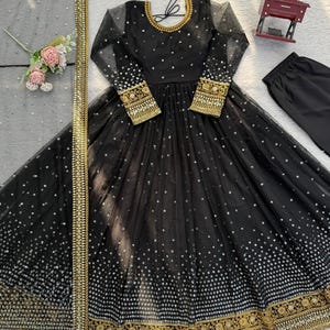 Peut inclure: Robe de soir&eacute;e noire &agrave; manches longues, orn&eacute;e de paillettes argent&eacute;es et de broderies dor&eacute;es. La robe pr&eacute;sente une jupe ample et est associ&eacute;e &agrave; un pantalon noir assorti et &agrave; une &eacute;charpe transparente &agrave; bordure dor&eacute;e. Un collier dor&eacute; et un accessoire floral pour les cheveux compl&egrave;tent l'ensemble.