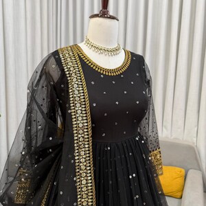 Peut inclure: Robe de soir&eacute;e noire orn&eacute;e de broderies dor&eacute;es et de sequins. La robe pr&eacute;sente une encolure ronde, des manches longues et une jupe fluide. Un ch&acirc;le noir transparent avec des d&eacute;tails de sequins et un collier dor&eacute; compl&egrave;tent l'ensemble.