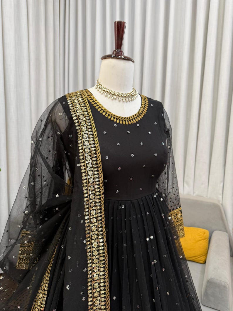 Peut inclure: Robe de soir&eacute;e noire orn&eacute;e de broderies dor&eacute;es et de sequins. La robe pr&eacute;sente une encolure ronde, des manches longues et une jupe fluide. Un ch&acirc;le noir transparent avec des d&eacute;tails de sequins et un collier dor&eacute; compl&egrave;tent l'ensemble.