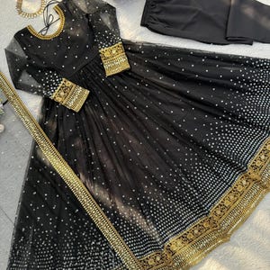 Peut inclure: Une robe de soir&eacute;e noire avec des broderies dor&eacute;es et des sequins. La robe a des manches longues, un col rond et une jupe fluide. Un pantalon noir assorti et une &eacute;charpe dor&eacute;e sont inclus.