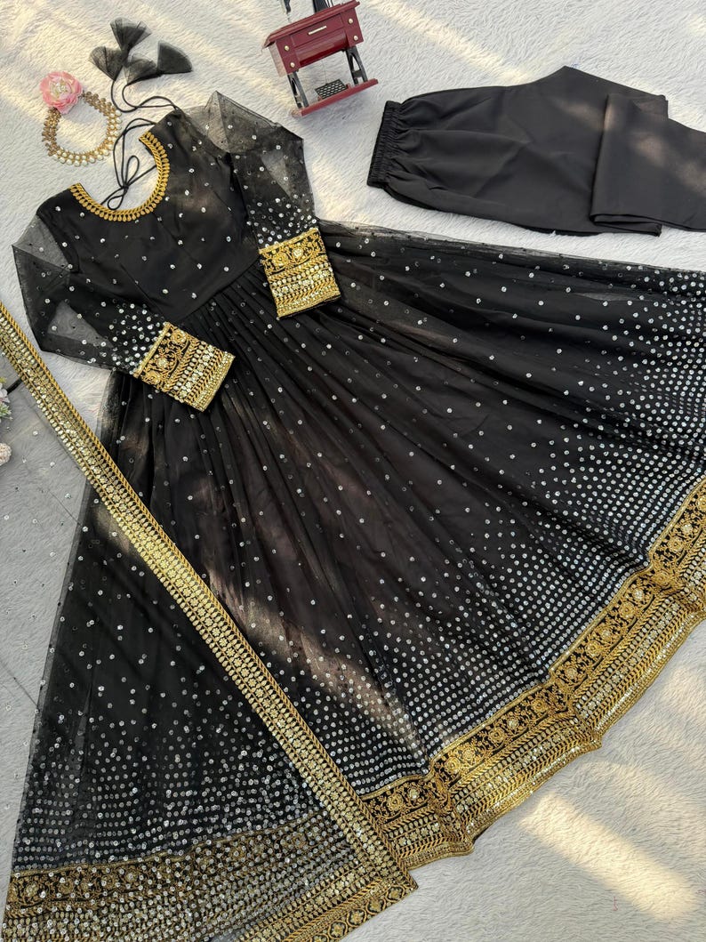 Peut inclure: Une robe de soir&eacute;e noire avec des broderies dor&eacute;es et des sequins. La robe a des manches longues, un col rond et une jupe fluide. Un pantalon noir assorti et une &eacute;charpe dor&eacute;e sont inclus.