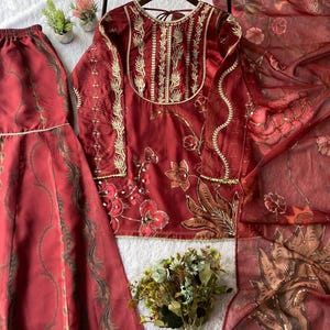 May include: A three-piece maroon outfit featuring a long-sleeved top with gold embroidery, matching skirt, and a sheer scarf. The top is hanging on a wooden hanger. Floral and leaf patterns adorn the fabric.