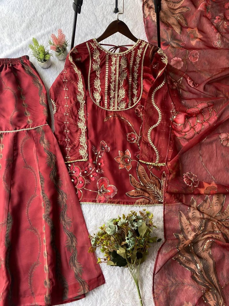May include: A three-piece maroon outfit featuring a long-sleeved top with gold embroidery, matching skirt, and a sheer scarf. The top is hanging on a wooden hanger. Floral and leaf patterns adorn the fabric.