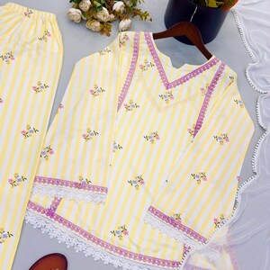 K&ouml;nnte beinhalten: Ein gelb-wei&szlig; gestreiftes Set aus einer lang&auml;rmeligen Tunika und einer passenden Hose mit kleinen Blumenmustern, violetter Spitzenborte am V-Ausschnitt und wei&szlig;er, bogenf&ouml;rmiger Spitzenkante an &Auml;rmeln und Saum.
