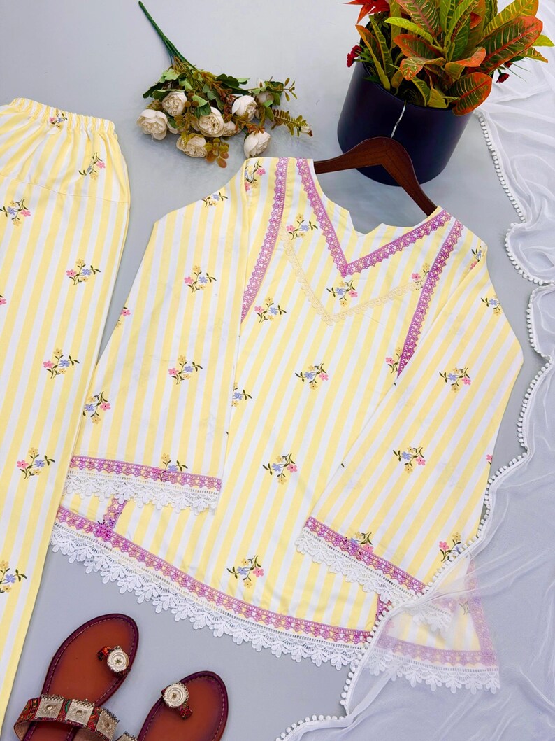 K&ouml;nnte beinhalten: Ein gelb-wei&szlig; gestreiftes Set aus einer lang&auml;rmeligen Tunika und einer passenden Hose mit kleinen Blumenmustern, violetter Spitzenborte am V-Ausschnitt und wei&szlig;er, bogenf&ouml;rmiger Spitzenkante an &Auml;rmeln und Saum.