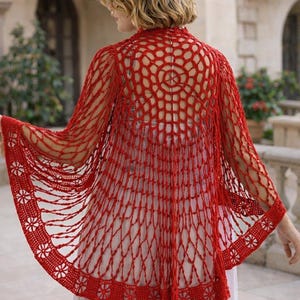 Puede incluir: Un chal de ganchillo rojo de tejido abierto con un patrón floral en los bordes. El chal presenta un diseño circular en la espalda y mangas largas. La prenda es ligera y parece hecha a mano.